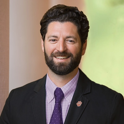 Portrait of a man with a beard, wearing a black suit and purple tie, smiling confidently.