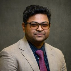 Professional headshot of a man wearing a beige suit and eyeglasses, with a dark shirt and pink tie.