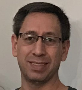 A smiling man with glasses and short hair against a light background.