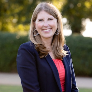A smiling woman with long blonde hair wearing a navy blazer and a coral top, standing outdoors.