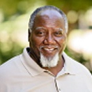 Smiling older man with a gray beard, wearing a beige polo shirt, set against a blurred green background.