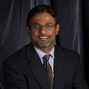 Professional portrait of a smiling man in a suit with glasses, set against a dark backdrop.
