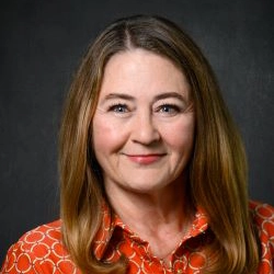 Professional headshot of a woman with long brown hair, wearing an orange patterned shirt.