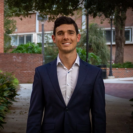 A young man in a suit smiles confidently while standing outdoors in an urban environment.