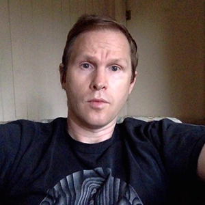A man with short hair is sitting on a couch, wearing a black T-shirt with a graphic design.