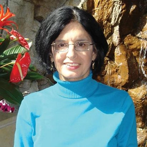 A woman in a blue turtleneck smiles in front of a water feature and tropical plants.