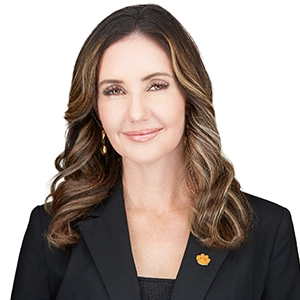Professional headshot of a woman with long, wavy hair wearing a black blazer and a subtle smile.
