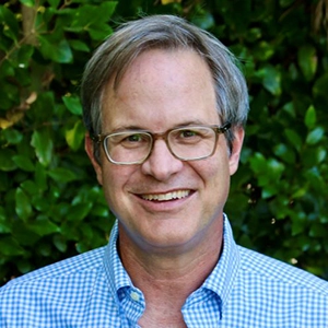 Portrait of a smiling man with glasses, wearing a blue checked shirt, set against a backdrop of green foliage.