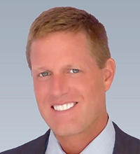 Professional headshot of a smiling man in a suit with short hair and light blue eyes.