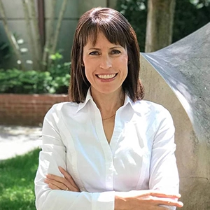 Professional portrait of a smiling woman in a white shirt, standing outdoors with arms crossed.