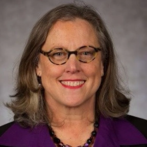 Professional headshot of a woman with gray hair and glasses, wearing a purple blouse and a necklace.