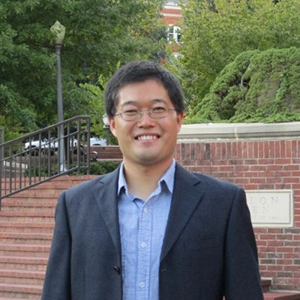 A smiling man in a blazer standing outdoors in front of a brick wall and greenery.