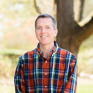 A smiling man wearing a plaid shirt stands outdoors in a natural setting.