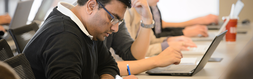 Male student with glasses writing with others in background on laptops.