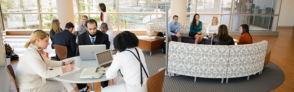 Many male and female students sitting at several tables with laptops and conversing with one another.