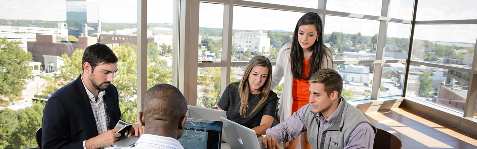 Three male and two female students around a table with laptops and city view in background.