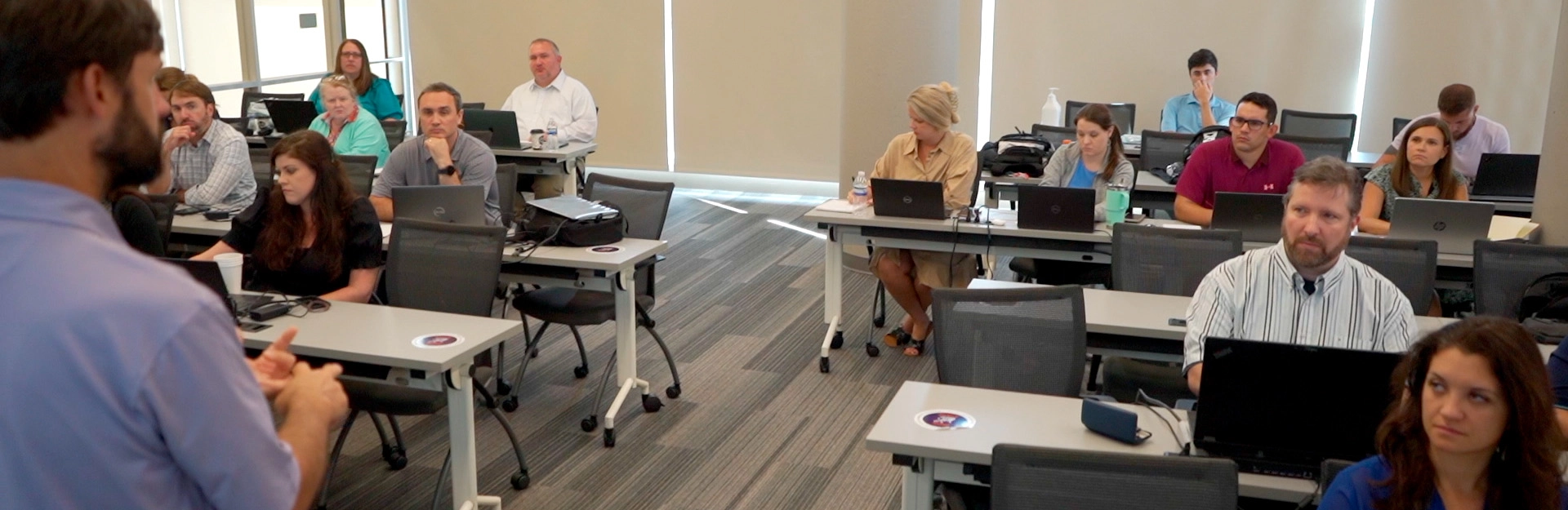 Many male and female students sitting at several tables with laptops in classroom environment..