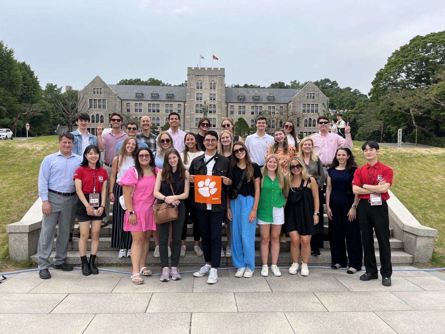 A group of diverse students poses in front of a historic building, holding a Clemson University sign.