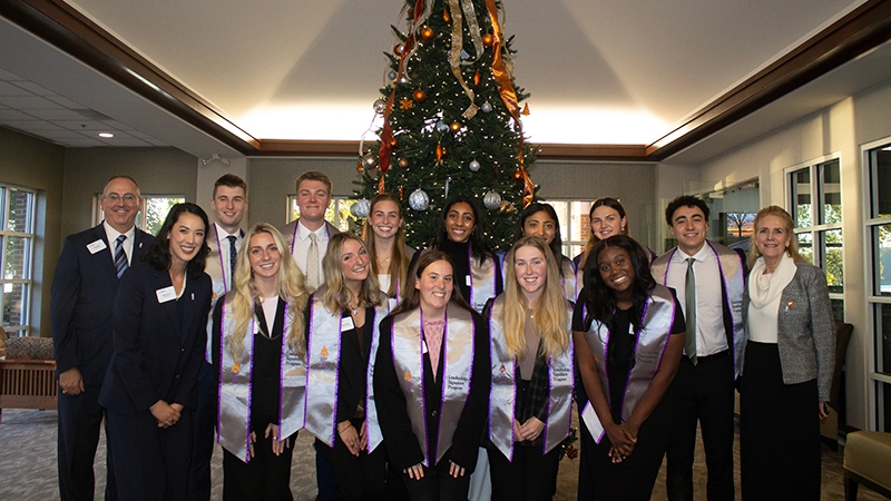 A group photo of students in graduation attire posing in front of a decorated Christmas tree, alongside faculty members.