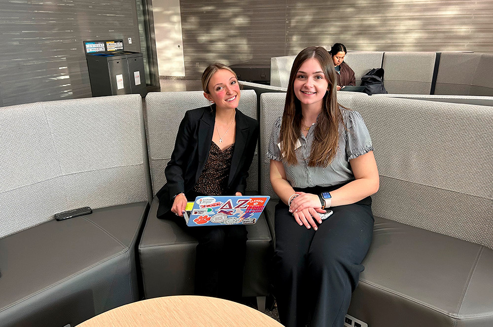 Two women sitting together on a modern couch, one using a laptop with colorful stickers while the other smiles at the camera.
