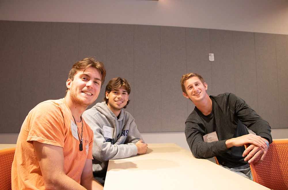 Three smiling young men sitting at a table in a casual setting.