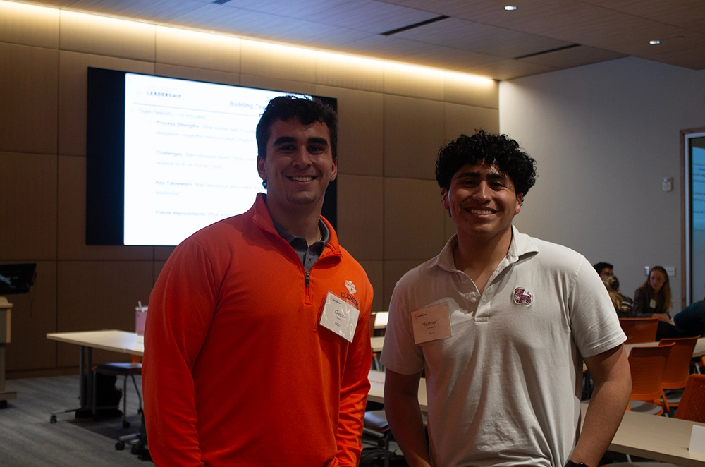 Two smiling young men standing in a conference room, with a presentation displayed in the background.