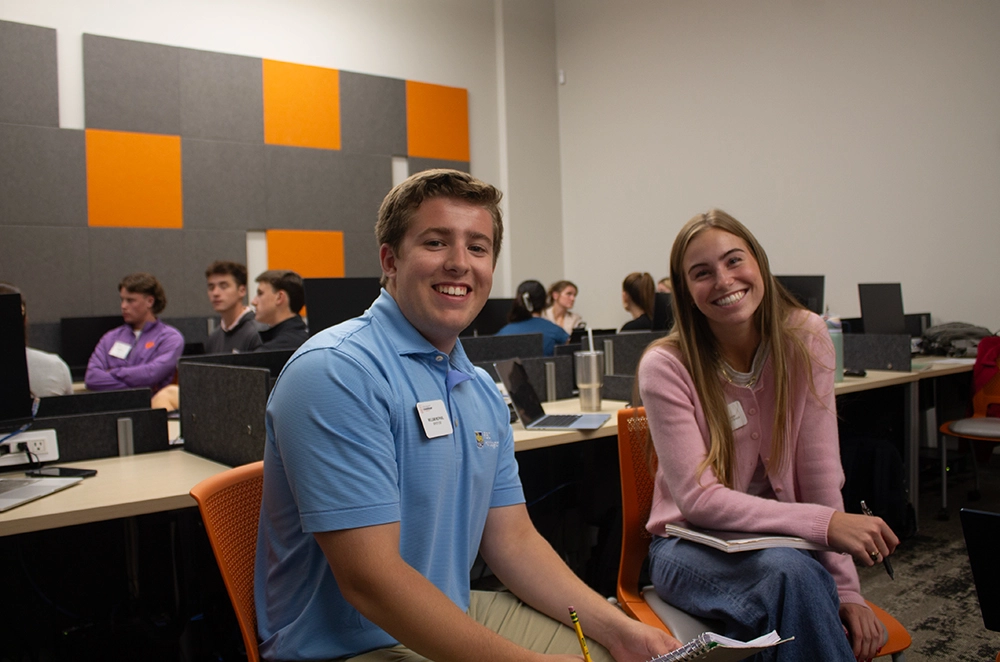 Two students smiling and engaging with each other in a classroom setting, with computers and classmates in the background.