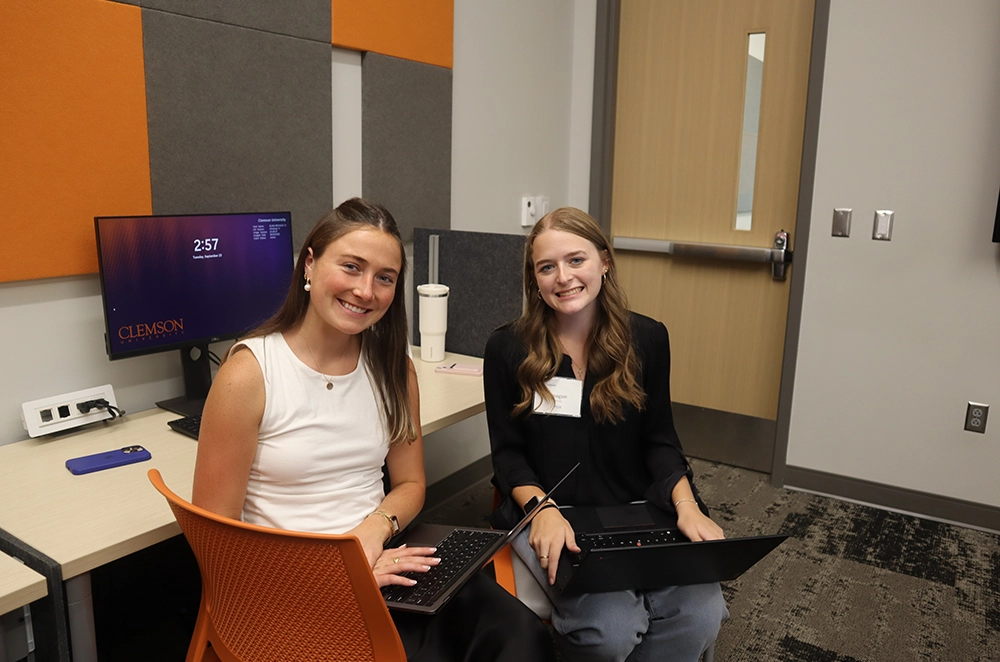 Two young women sitting in a modern workspace, each with a laptop in front of them, smiling and engaged in conversation.