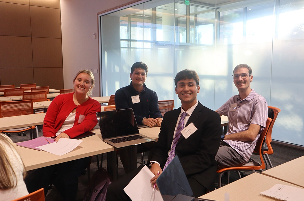 A group of four students smiling and sitting around a table in a classroom setting, engaged in a collaborative activity.