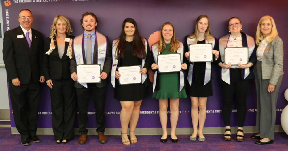 A group of six graduates posing with their certificates, dressed in formal attire, in front of a university backdrop.