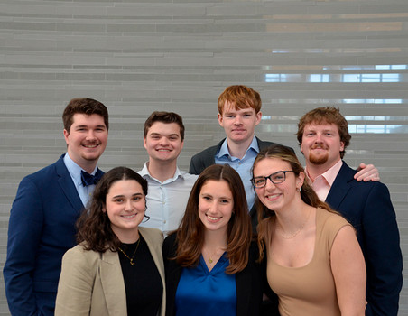A group photo of seven young professionals in business attire, smiling and posing together in a modern setting.