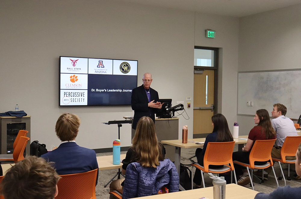 Presenter speaks to a seated audience in a classroom beside a screen displaying “Dr. Buyer’s Leadership Journey” and university logos.