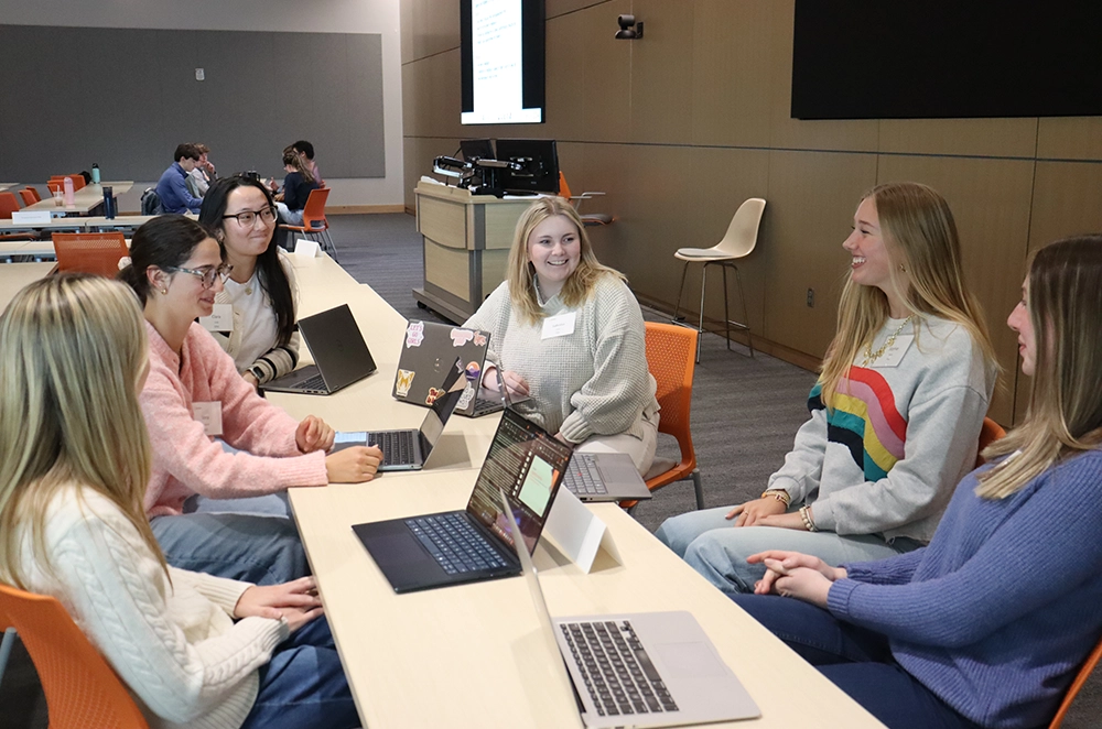 Students collaborate around tables with laptops during a workshop in a classroom.
