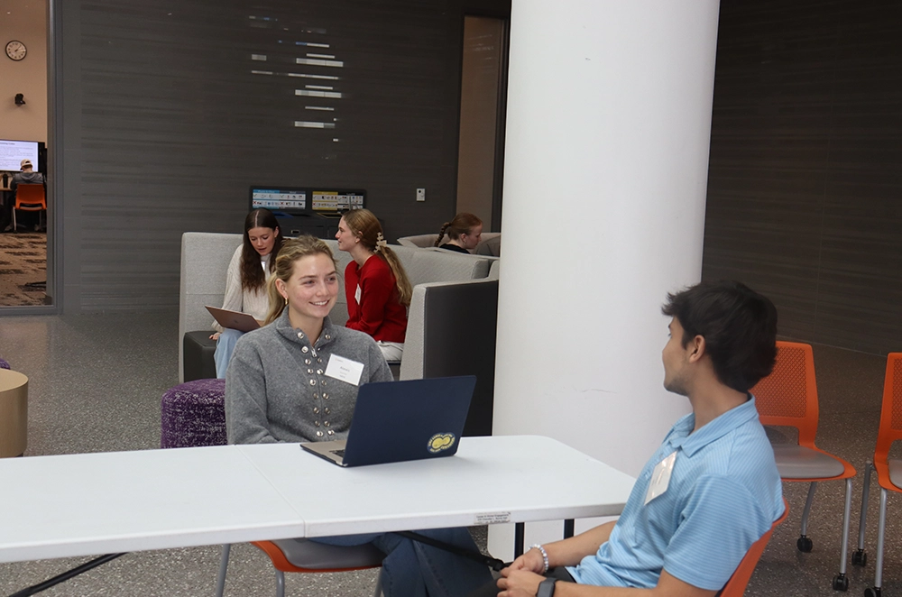 Two students talk across a table with a laptop in a campus lounge while other students study in the background.