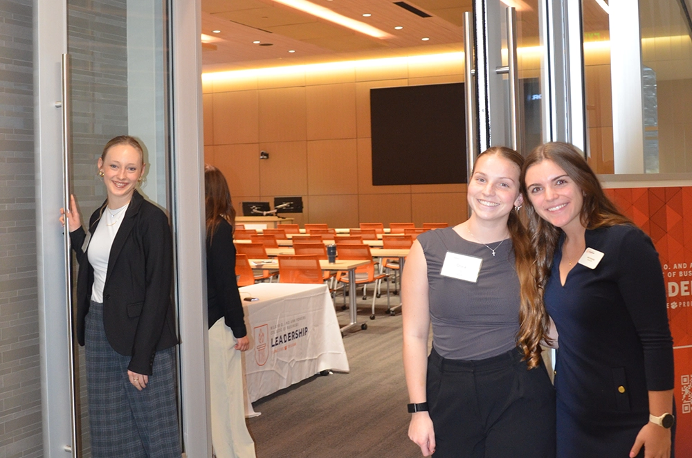Three attendees smiling at the entrance to a leadership event in a modern conference room with rows of orange chairs.