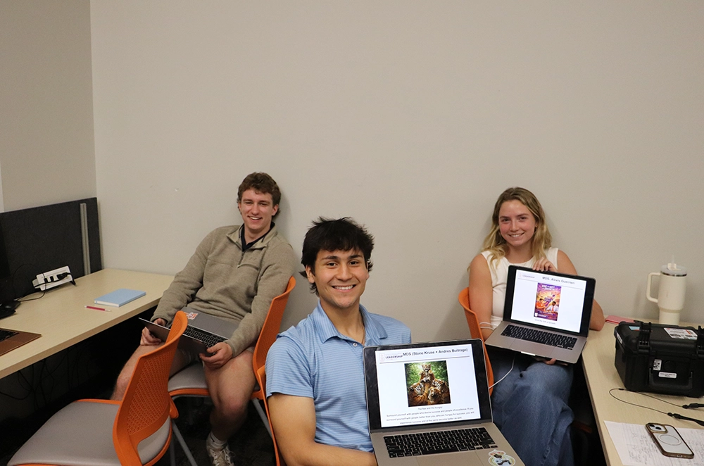 Three students in a study room smile while holding open laptops displaying presentation slides.