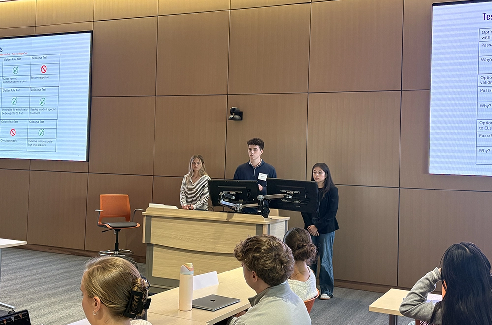Three students give a presentation at the front of a lecture hall with slides displayed on large screens.