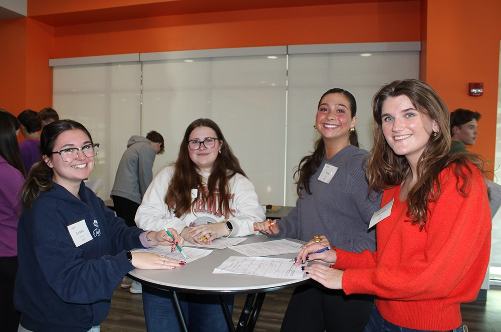 Four students smiling while working together at a round table with papers and pens.