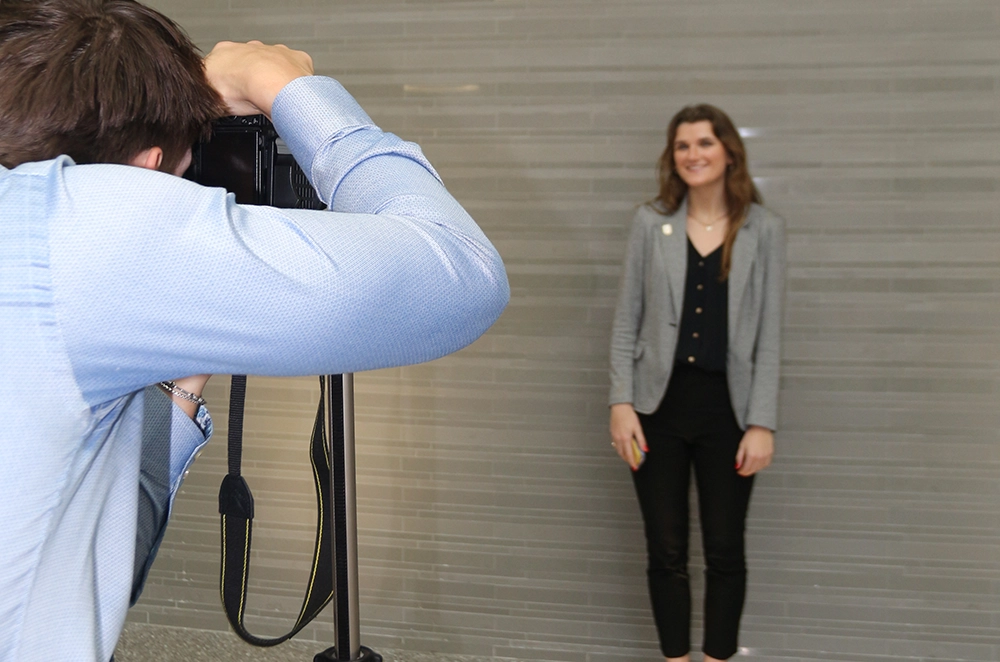 Photographer taking a portrait of a woman standing against a gray wall.