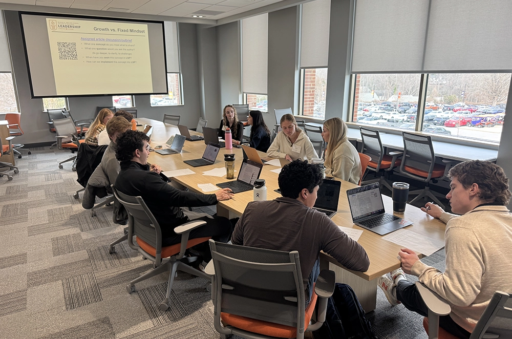 Students collaborate around a conference table with laptops during a workshop, with a “Growth vs. Fixed Mindset” slide projected on the screen.