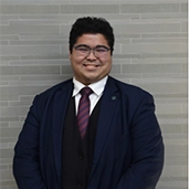 Person in a navy suit and striped tie smiling in front of a light gray brick wall.