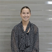 Smiling person in a dark patterned blouse standing in front of a light gray tiled wall.
