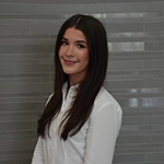 Professional headshot of a young woman with long dark hair, wearing a white button-up shirt against a light gray tiled background.