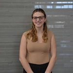 A smiling young woman with long, wavy hair wearing glasses and a beige top stands in front of a gray background.