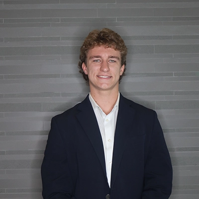 Portrait of a young man in a suit and collared shirt with a friendly smile against a modern gray background.
