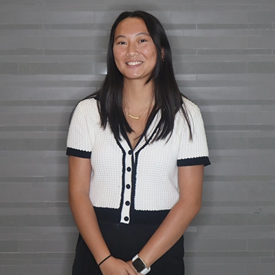 A smiling young woman wearing a black and white cardigan stands against a gray tiled backdrop.