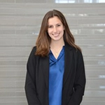 A smiling woman wearing a blue blouse and black blazer stands in front of a modern backdrop.