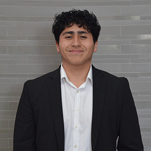 Man with short dark curly hair wearing a dark jacket and white shirt and smiling.
