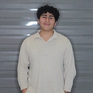 A young man with curly hair smiles while wearing a light-colored shirt against a gray background.