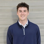 Smiling young man in a navy quarter-zip sweater standing against a light brick wall.
